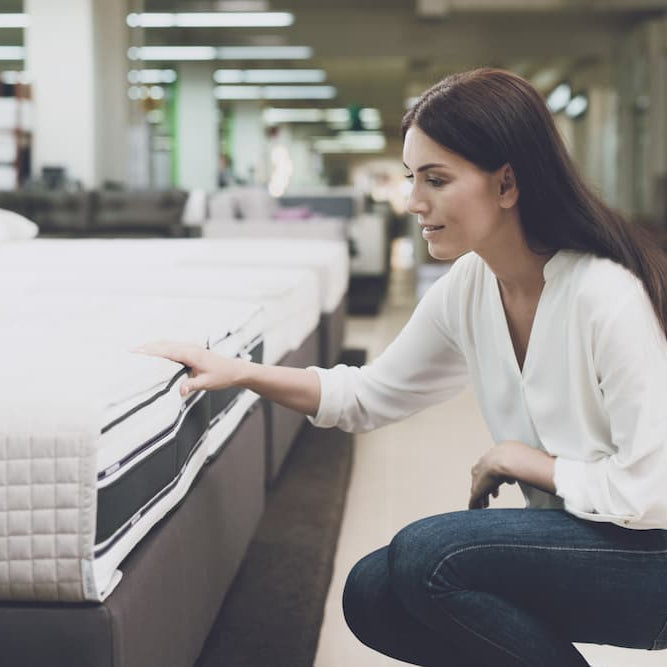 A woman in a white shirt and jeans in a mattress store. She examines the mattress she wants to buy.